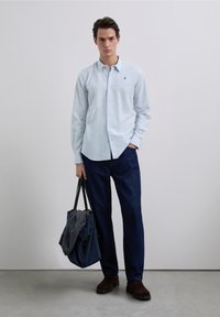 Lichtblauw overhemd met knopen en een subtiel ruitpatroon, gecombineerd met donkerblauwe broek en bruine schoenen. Houdt een donkerblauwe tas vast.