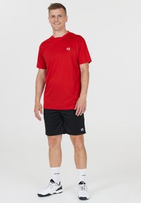 Rød sports t-shirt med rund hals, parret med sorte shorts. Har logo på brystet og shortsene. Skoene er hvide med sorte detaljer.