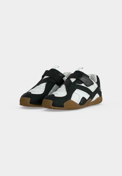 Paire de chaussures de sport blanches et noires avec semelles marron et brides croisées, présentées sur un fond gris clair uni.