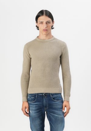 Beige gebreide trui met een textuurpatroon, ronde halslijn en geribbelde boorden en zoom. Gepaard met donkerblauwe versleten jeans.