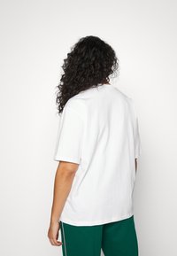 Valkoinen puuvillapaita, jossa on rento leikkaus, lyhyet hihat ja pyöreä pääntie, nähtynä takaa, yhdistettynä vihreisiin housuihin.