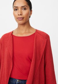 Rote Strickjacke mit offenem Vorderteil und langen Ärmeln, über einem roten kurzärmligen Oberteil getragen. Glatte Textur, lässiges Design, minimale Details.