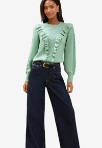 Pulover mint green cu un design șifonat pe de-a lungul gulerului și mânecilor, asociat cu blugi evazați din denim închis la culoare, cu talie înaltă.