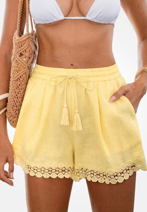 Kobieta ubrana w żółte lniane szorty z falbanką z koronki, biały top od bikini, trzymająca tkany jasnobrązowy worek na jednym ramieniu.