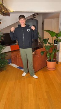 Sweat à capuche noir côtelé avec fermeture éclair et logo sur la poitrine, accompagné d'un pantalon de survêtement vert et de baskets blanches. Intérieur avec plantes et mobilier.