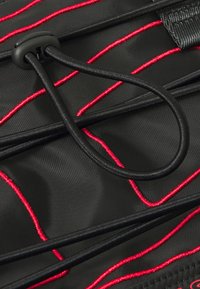 Tissu noir avec des coutures rouges ; comprend des cordons élastiques et un cordon de serrage. Surface texturée avec des lignes et une zone renforcée pour la durabilité.