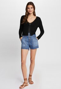 Cardigan nero a coste con scollo a V, indossato sopra una maglia scura, abbinato a pantaloni corti di jeans blu a vita alta con chiusura a bottone e tasche.