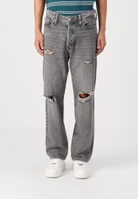 Jeans de mezclilla gris desgastados con grandes desgarros en las rodillas, corte recto y un acabado deslavado. Combinados con zapatillas blancas.