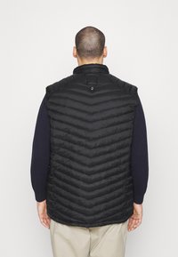 Gilet nero imbottito con motivo a rombi, colletto alto e chiusura posteriore con bottoni. Indossato sopra una camicia a maniche lunghe blu navy.