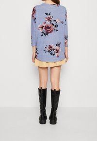 Blommigt mönstrad, långärmad blå topp med rundad nederkant, bärs över en gul kjol och svarta knähöga stövlar. Slät textur.