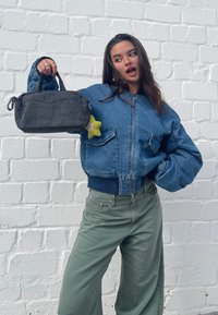 Džinsinė bombonešio striukė su banguotais rankogaliais, derinama su šviesiai žaliais plačiomis kelnėmis. Modelis laiko tamsiai pilką krepšį su žvaigždės formos raktų pakabuku.