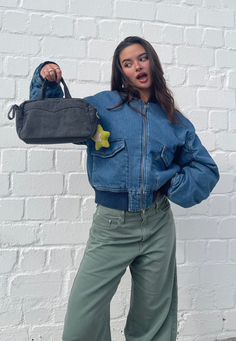 Jachetă bomber din denim cu manșete striate, asortată cu pantaloni largi de culoare verde deschis. Modelul ține o geantă gri închis cu un breloc în formă de stea.