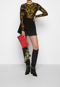 Czarny bodysuit z długim rękawem zdobiony złotymi wzorami paisley, w zestawieniu z czarną plisowaną mini spódnicą, czarnymi kozakami do kolan i czerwoną torebką kopertową.