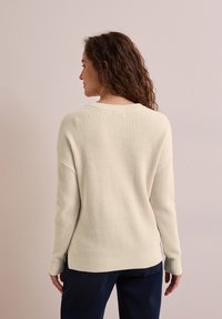 Hellbeiger Strickpullover mit geripptem Muster, lässiger Schnitt, Rundhalsausschnitt und langen Ärmeln, kombiniert mit dunkelblauen Hosen.