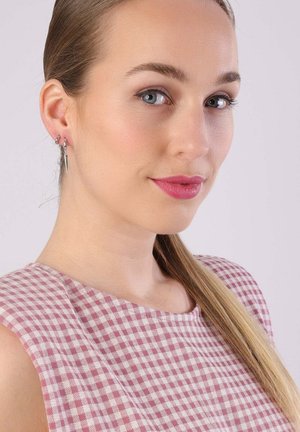 Boucles d'oreilles en argent avec un design pointu, portées par un modèle aux cheveux droits et clairs, dans un haut sans manches à carreaux roses et blancs.