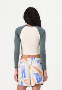 Top corto con corpo beige e maniche lunghe color teal; abbinato a pantaloni corti multicolori con motivi astratti in blu, arancio e verde.