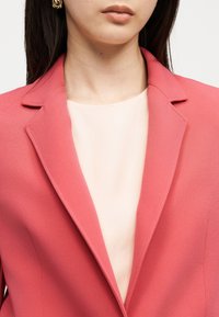 Γυναίκα που φορά χρυσούς κρίκους στα αυτιά και ένα κοραλλί σακάκι πάνω από ανοιχτό μπεζ τοπ, εμφανιζόμενη από τους ώμους έως τα χείλη μπροστά σε απλό φόντο.