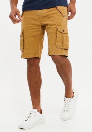 Shorts cargo kaki avec plusieurs poches, une coupe décontractée et une texture lisse, associés à des baskets blanches au design minimaliste.