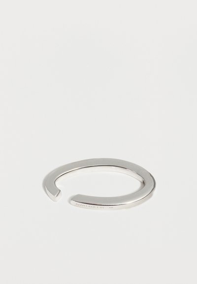 Bracelet manchette en argent ouvert avec une finition lisse et un nom de marque subtilement gravé sur un côté, présenté sur un fond blanc.