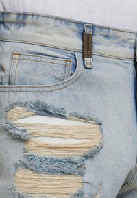 Šviesiai mėlyni nusidėvėję džinsai su suplėšytais ir nugrandytais raštais, vario spalvos siūlais ir metaliniu ženkleliu ant juosmens.