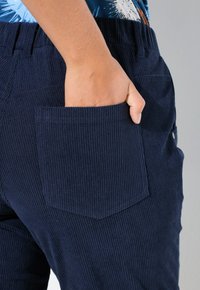 Mörkblå kordflortbyxor med en texturerad yta, med en stor bakficka och en hand som vilar inuti, elastisk midja synlig.