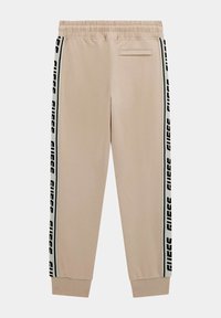 Beige sweatpants met een elastische tailleband, voorzien van zwart-witte "GUESS" logo zijstrips en een achterzak. Voorzien van een gladde stof en een taps toelopende pijp.