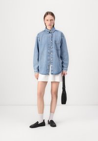Camicia di jeans azzurra chiara con bottoni e tasca sul petto, indossata sopra un vestito bianco. Scarpe nere e calzini grigi completano l'outfit.