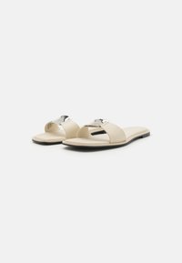 Beige slide sandals featuring a flat sole, open toe design, and a metallic accent on the upper strap. Smooth texture overall.