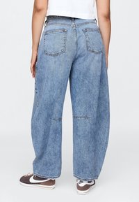 Vaqueros de mezclilla azul claro, holgados, de cintura alta, con grandes bolsillos traseros y detalles de dobladillo sin rematar. Combinados con zapatillas marrones y blancas.