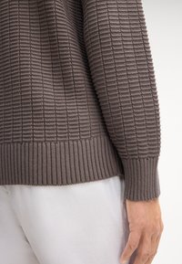 Sweter z teksturowanego dzianiny w stonowanej brązowej odsłonie, z prążkowanymi mankietami i dołem. Posiada wzór w kratę, który dodaje głębi. W połączeniu z białymi spodniami.