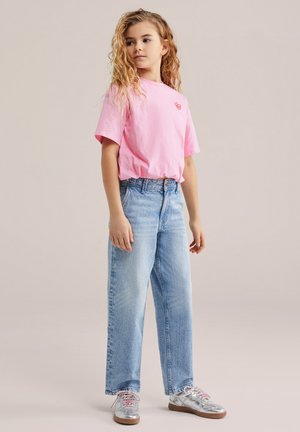 Fiatal lány hosszú, göndör hajjal, rózsaszín pólóban, világoskék farmerben és fémes ezüst sportcipőben, semleges háttér előtt állva.