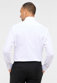 Weißes langärmliges Hemd mit Standardkragen; glatte Textur; schmaler Schnitt; getragen mit dunklen Hosen; Rückansicht zeigt Knopfmanschetten und Taschen.