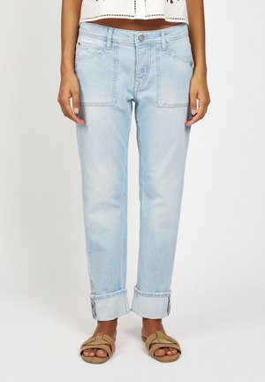 Frau trägt hellblaue, umgeschlagene Jeans mit vorderen aufgesetzten Taschen und braunen Sandalen mit offenem Zehenbereich, stehend vor einem schlichten Hintergrund.