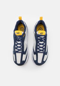 Pár bílých a námořnických tenisek Nike se žlutými akcenty, námořnickými tkaničkami a květinovým vzorem viditelným na žlutých vložkách, z pohledu shora.