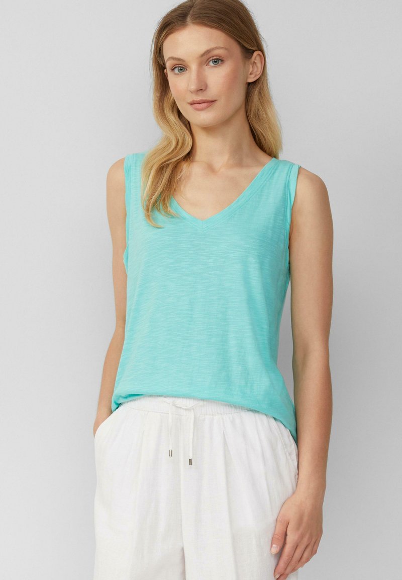 Een mouwloze, V-hals tanktop in een lichte aqua kleur, gemaakt van een zachte, gestructureerde stof. In combinatie met losse, witte linnen broeken.