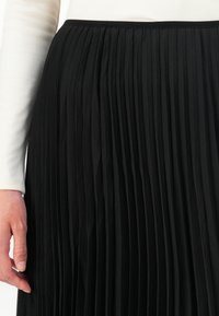 Mão descansando ao lado de uma saia preta com pregas, vestindo uma blusa de manga longa branca com nervuras, close-up da cintura até a metade da coxa.