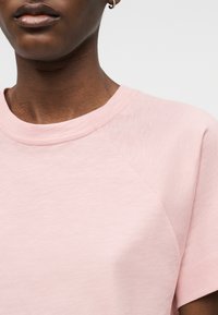 Κοντινό πλάνο ενός ατόμου που φοράει ένα ανοιχτό ροζ μπλουζάκι με στρογγυλή λαιμόκοψη και ένα μικρό σκουλαρίκι κρίκο, με φόντο ένα απλό φόντο.