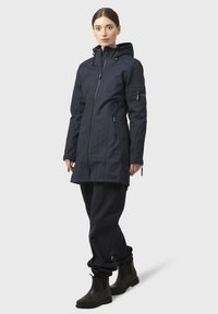 Chaqueta impermeable azul marino con capucha, cremallera frontal y múltiples bolsillos; combinada con pantalones oscuros y botas negras de tobillo.