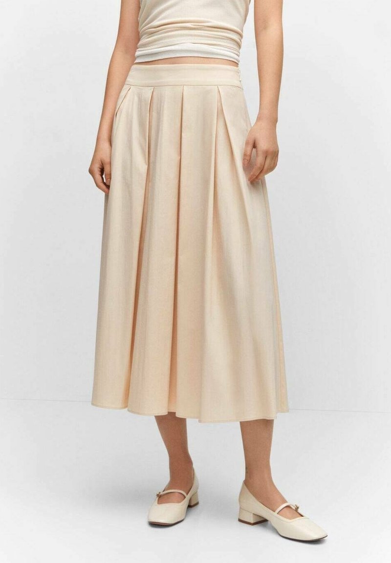 Femme portant une jupe midi plissée beige, un top sans manches beige et des chaussures Mary Jane crème à talons bas, debout les mains détendues.