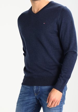 Homme portant un pull bleu marine à col en V et un jean bleu, la main droite reposant près de sa poche.