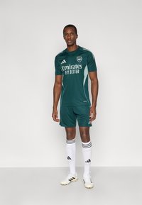 Grønt fodboldtøj med rund hals, der har hvide accenter og tekst. Bruges med hvide sokker, sort-hvide fodboldstøvler og atletisk kropsbygning.