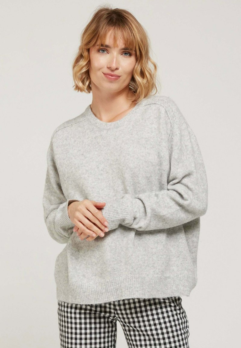 Pull en tricot gris avec un col rond et des poignets côtelés, associé à un pantalon à motif vichy noir et blanc. Texture douce et coupe décontractée.