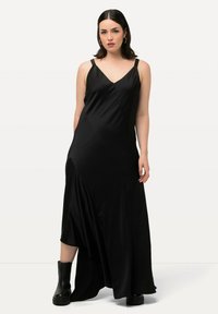 Robe maxi noire avec un décolleté en V profond, des bretelles fines et un ourlet asymétrique. Fabriquée dans un matériau semblable à du satin avec une texture lisse. Portée avec des bottines.