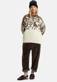 Bruin en crèmekleurig gebreid trui, corduroy bruine broek, zwarte beanie, en geruite slide sandalen met sokken, wat een gelaagd casual outfit creëert.