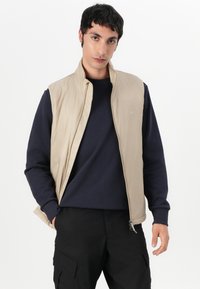 Beige isolierte Weste mit Reißverschluss vorne, hohem Kragen und navy-blauem Langarmshirt darunter. Getragen mit schwarzen Cargo-Hosen. Glattes, leichtes Material.