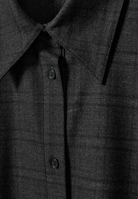 Gros plan d'une chemise boutonnée gris foncé montrant le col, la texture du tissu, un motif à petits carreaux et trois boutons noirs visibles attachés.