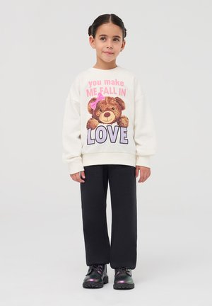 Felpa bianca con grafica di un orso e testo, abbinata a pantaloni neri e scarpe scure. La felpa presenta dettagli rosa e una vestibilità oversize.