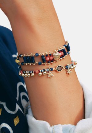 Bracelet multi-brins présentant des perles bleu marine et corail, des accents dorés, avec des charms en forme d'étoile et de goutte d'eau, disposés dans un design superposé.