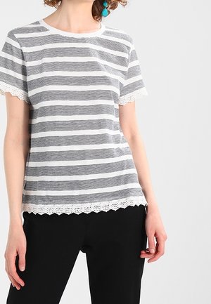 Kvinde iført en grå og hvid stribet kortærmet top med blondedetaljer på ærmer og kant, kombineret med sorte bukser og turkise øreringe.