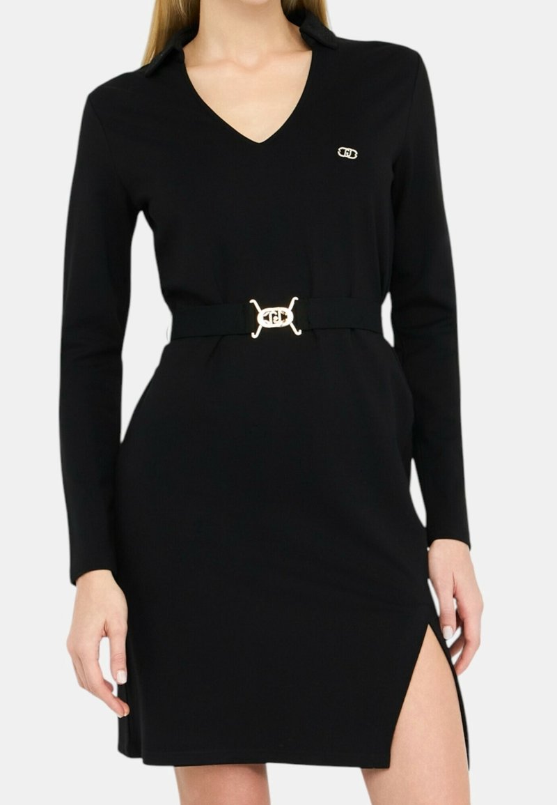 Femme portant une robe noire à manches longues avec un col en V, une ceinture à boucle dorée, un petit logo sur la poitrine et une fente haute à droite.